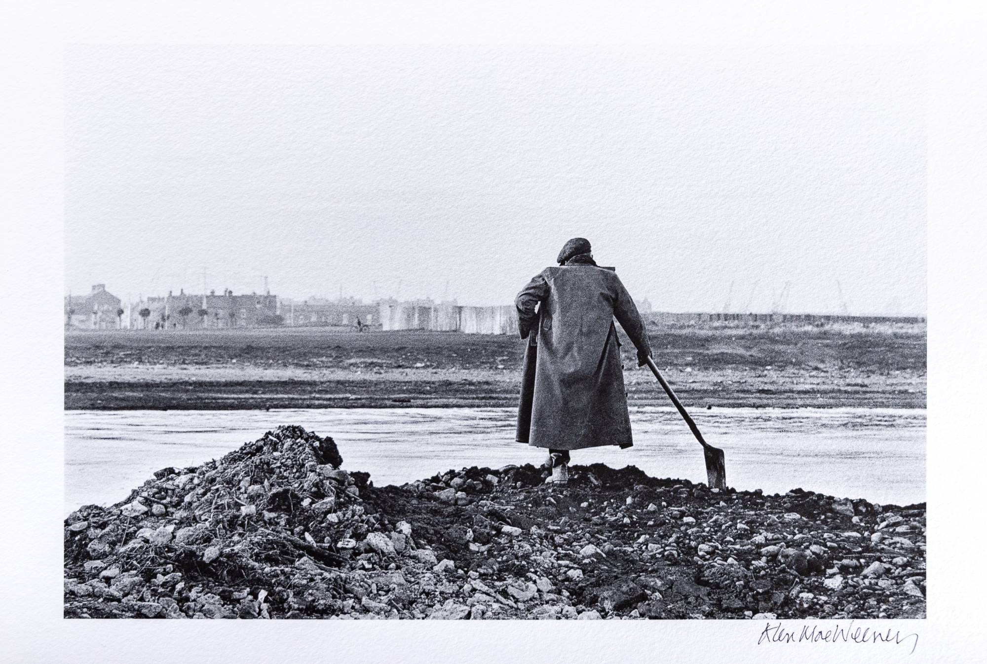 man standing on rocky river bank leaning on shovel looking out across the river